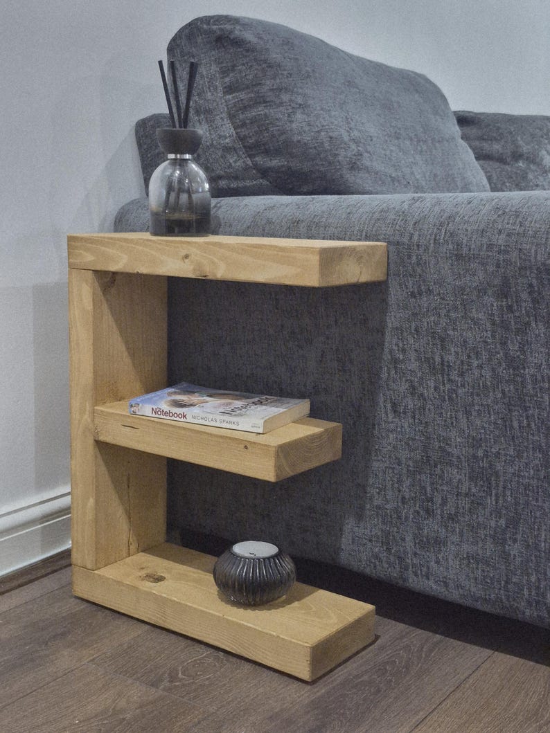 May include: A light-colored wooden side table with two shelves. A book, a candle holder, and a diffuser bottle with reeds are on the shelves. The table is next to a gray sofa.