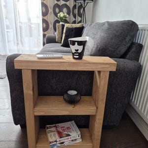 May include: A light brown wooden side table with two shelves. A black and white face-shaped mug sits on top, along with a remote. The shelves hold a small black candle holder and two books. The table is next to a grey sofa.