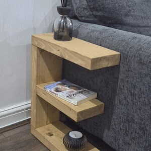 May include: A light-colored wooden side table with three shelves. A clear glass diffuser bottle with black reeds sits on the top shelf. A book titled "Notebook" rests on the middle shelf, and a small candle holder is on the bottom shelf.