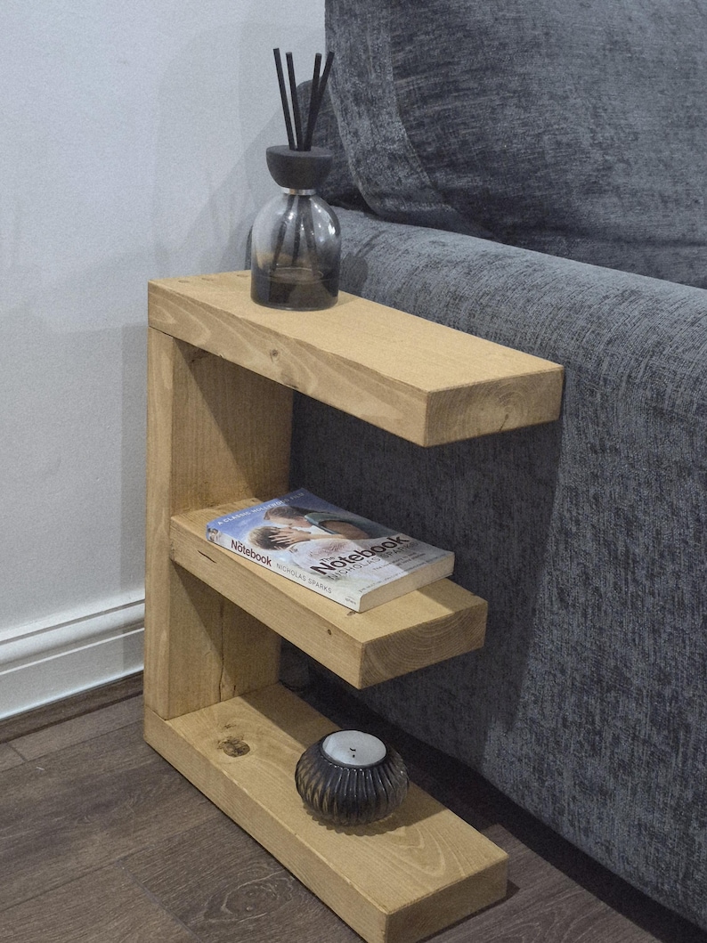 May include: A light-colored wooden side table with three shelves. A clear glass diffuser bottle with black reeds sits on the top shelf. A book titled "Notebook" rests on the middle shelf, and a small candle holder is on the bottom shelf.
