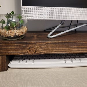 May include: A dark brown wooden monitor stand with a keyboard underneath. A glass terrarium with small succulents and pebbles sits on top. The stand is designed to elevate a computer monitor.
