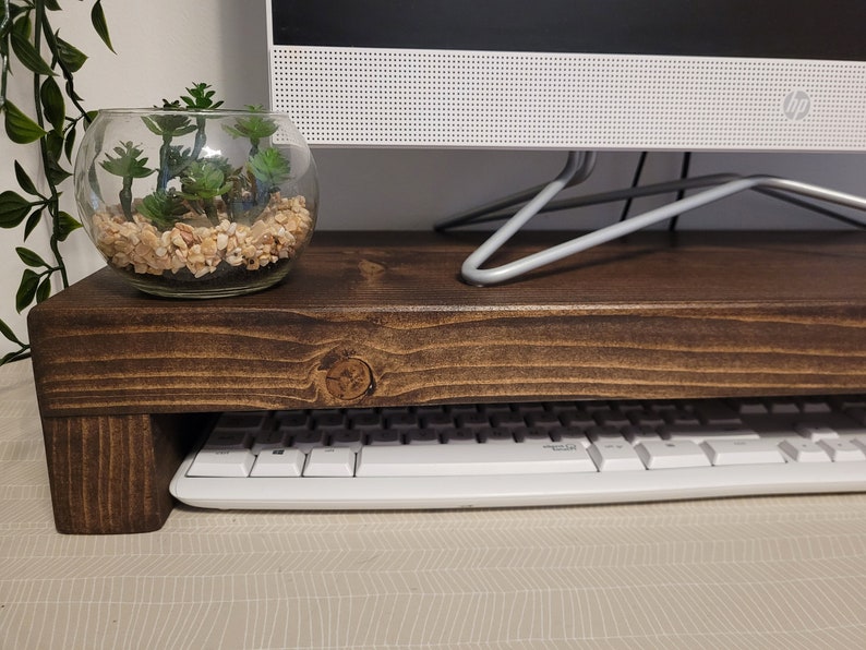 May include: A dark brown wooden monitor stand with a keyboard underneath. A glass terrarium with small succulents and pebbles sits on top. The stand is designed to elevate a computer monitor.