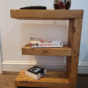 May include: A three-tiered, wooden side table with a warm, natural finish. The table holds a remote control, a glass bowl with decorative elements, a book titled "Invited Her In", and a small, white box with a colorful stack of cards.