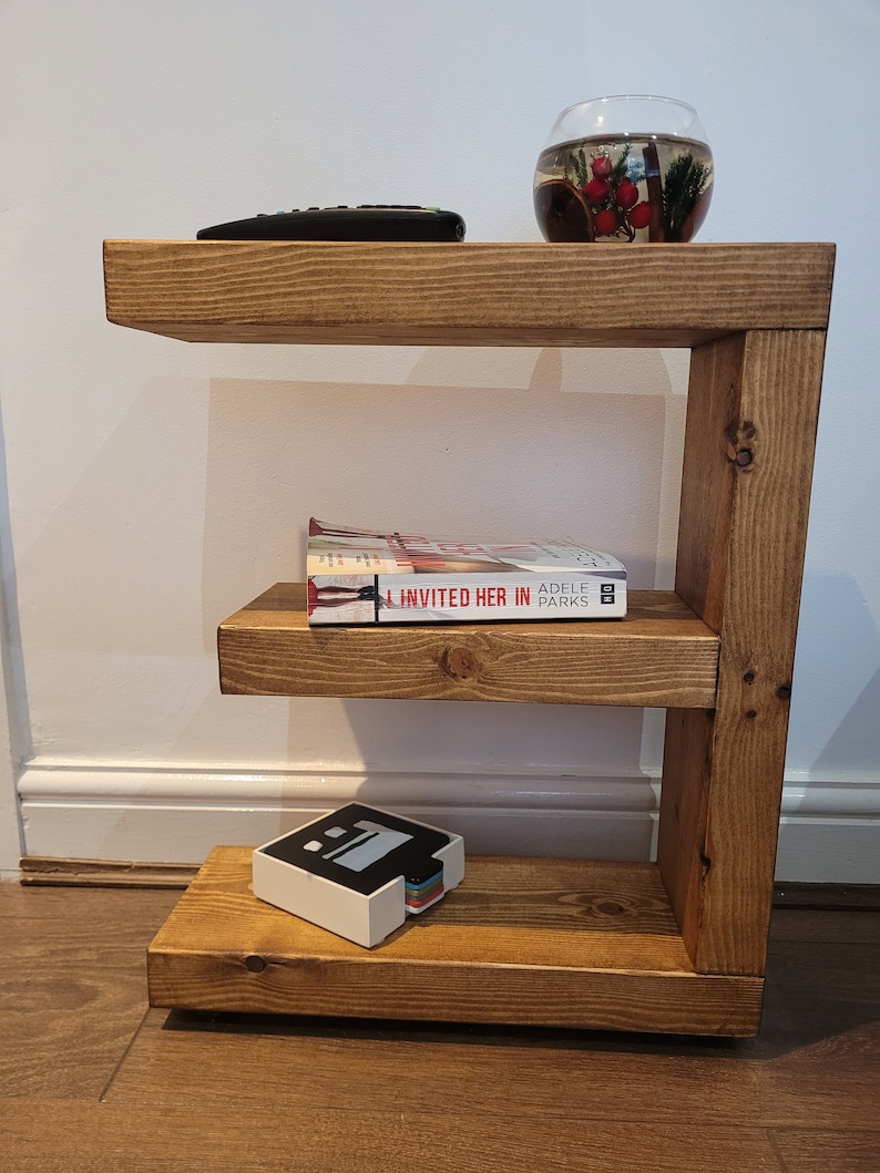 May include: A three-tiered, wooden side table with a warm, natural finish. The table holds a remote control, a glass bowl with decorative elements, a book titled "Invited Her In", and a small, white box with a colorful stack of cards.