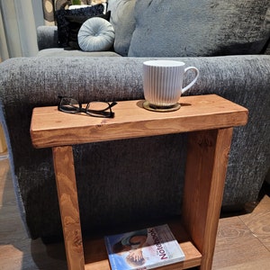 May include: A small, stained wooden side table with a lower shelf. A pair of black glasses, a white mug with vertical lines, and a coaster sit on top. A copy of "Notebook" by Nicholas Sparks rests on the shelf. The table is next to a grey sofa.