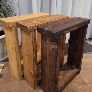 May include: Three wooden side tables in varying shades of brown. The tables have a rectangular design with a top and a lower shelf, and are approximately 61 cm tall. The wood grain is visible, adding to the rustic aesthetic.