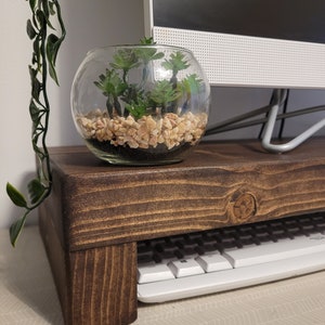 May include: A dark brown wooden monitor stand with a keyboard shelf. A clear glass bowl with small green plants and pebbles sits on top. A white computer monitor is in the background.