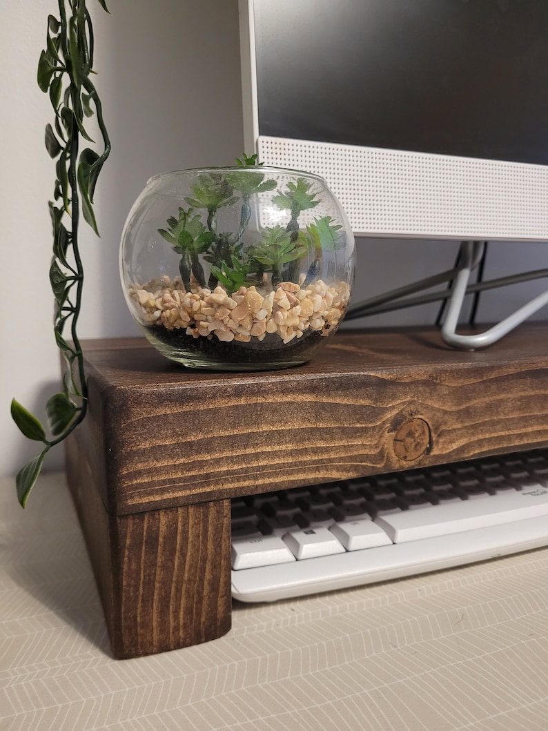 May include: A dark brown wooden monitor stand with a keyboard shelf. A clear glass bowl with small green plants and pebbles sits on top. A white computer monitor is in the background.