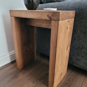May include: A small, handcrafted wooden side table with a warm, brown finish. The table has a rectangular top and angled legs, creating a modern, minimalist design. A dark bowl and a small white dish sit on top. The table is approximately 50 cm tall.