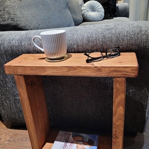 May include: A rectangular wooden side table with a medium brown finish, featuring a top surface holding a white ribbed ceramic mug and black-framed eyeglasses, and a lower shelf containing a book titled The Notebook.