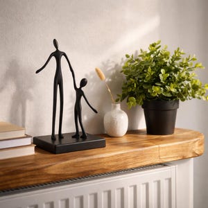 May include: A matte black sculpture depicting two stylised figures of different heights standing together on a rectangular base. The taller figure holds the hand of the smaller figure, both featuring elongated limbs and smooth, minimalist forms.