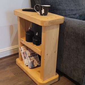 May include: A wooden side table with two shelves. The top shelf has a black mug and a remote control. The bottom shelf has two black candles and three books. The books are titled "Fractured" and "I Invited Her In".