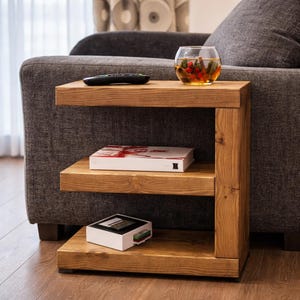 May include: A three-tiered, solid wood side table with a natural finish. The table has a rectangular top and shelves, with a vertical support on one side. A remote, a glass bowl with decorative items, and two boxes are on the shelves.