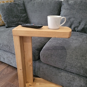May include: A wooden C-shaped side table with a rectangular top. A remote control and a white mug with a heart are on the table. The table is next to a grey sofa. The table's height is approximately 60 cm.