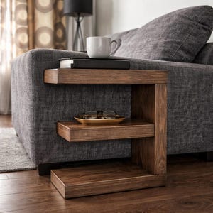May include: A dark brown wooden side table with three shelves. A white mug and two books sit on the top shelf. A small, gold-colored tray with eyeglasses rests on the middle shelf. The table is next to a gray sofa.