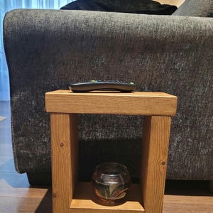 May include: A small, rectangular wooden side table with a natural light brown finish and visible wood grain. The table features a flat top surface, two vertical side supports, and a lower shelf holding a small, clear, round glass bowl.