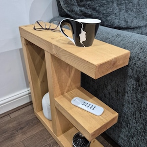 May include: A wooden side table with two shelves, holding a black and white mug, glasses, a remote, and decorative items. The table is made of light-coloured wood and is positioned next to a grey sofa.