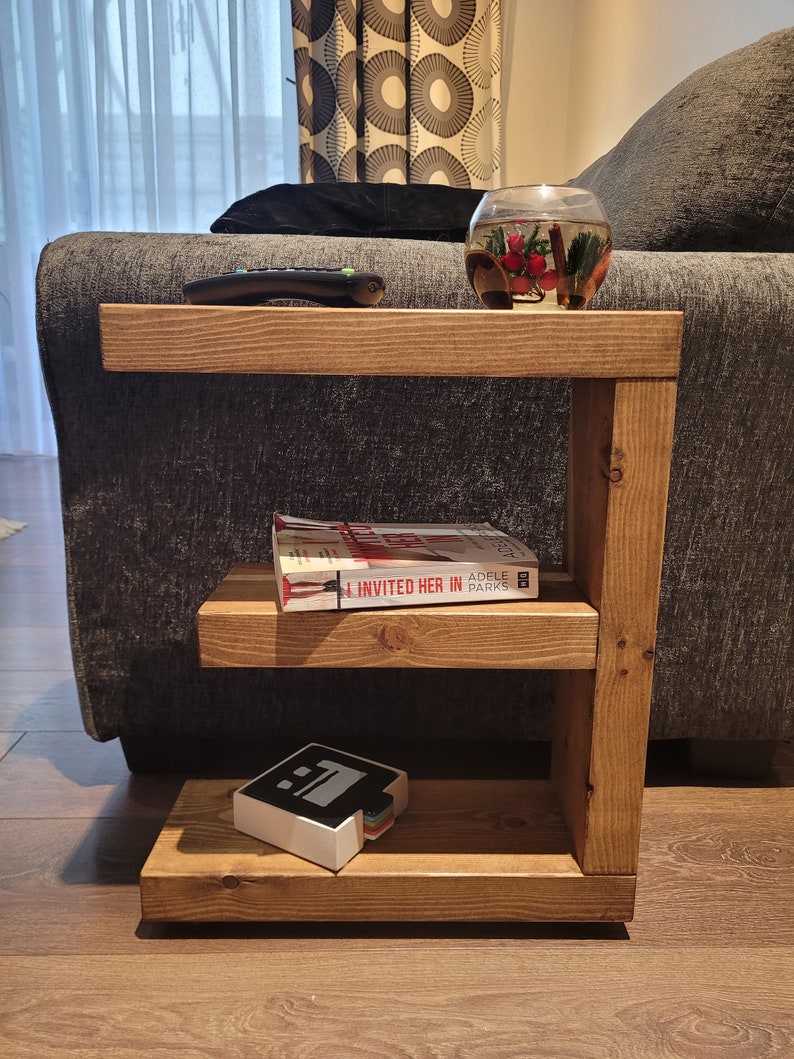 May include: A wooden side table with three shelves. The top shelf holds a remote and a glass bowl with decorative items. The middle shelf has a book titled "I Invited Her In". The bottom shelf has a white and black object.