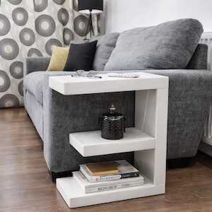 May include: A white side table with three shelves is positioned next to a gray sofa. A black decorative jar sits on the middle shelf, and books are stacked on the bottom shelf. The table is made of wood and has a modern design.