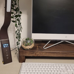 May include: A brown desk lamp with a digital display showing the time, a white keyboard, and a computer monitor on a wooden stand. A small glass terrarium with plants sits on the stand. Green vines hang from above.