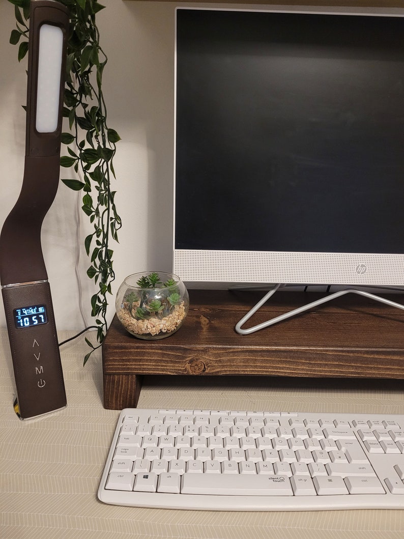 May include: A brown desk lamp with a digital display showing the time, a white keyboard, and a computer monitor on a wooden stand. A small glass terrarium with plants sits on the stand. Green vines hang from above.