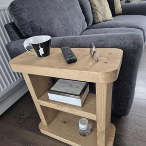 May include: A light-toned wooden side table with a rectangular top and a lower shelf. The table features rounded corners and vertical support legs. On the top surface sits a black and white mug, a black remote control, and a pair of eyeglasses.