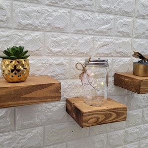May include: Three wooden shelves mounted on a white brick wall. The top shelf has a gold pineapple planter with a succulent plant. The middle shelf has a glass jar with lights inside. The bottom shelf has a gold butterfly figurine on a small box.