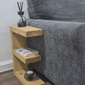 May include: A light-colored wooden side table with two shelves, positioned next to a gray sofa. The top shelf holds a glass diffuser with black reeds. A book and a small candle holder are on the lower shelves. The table is a functional piece of furniture.