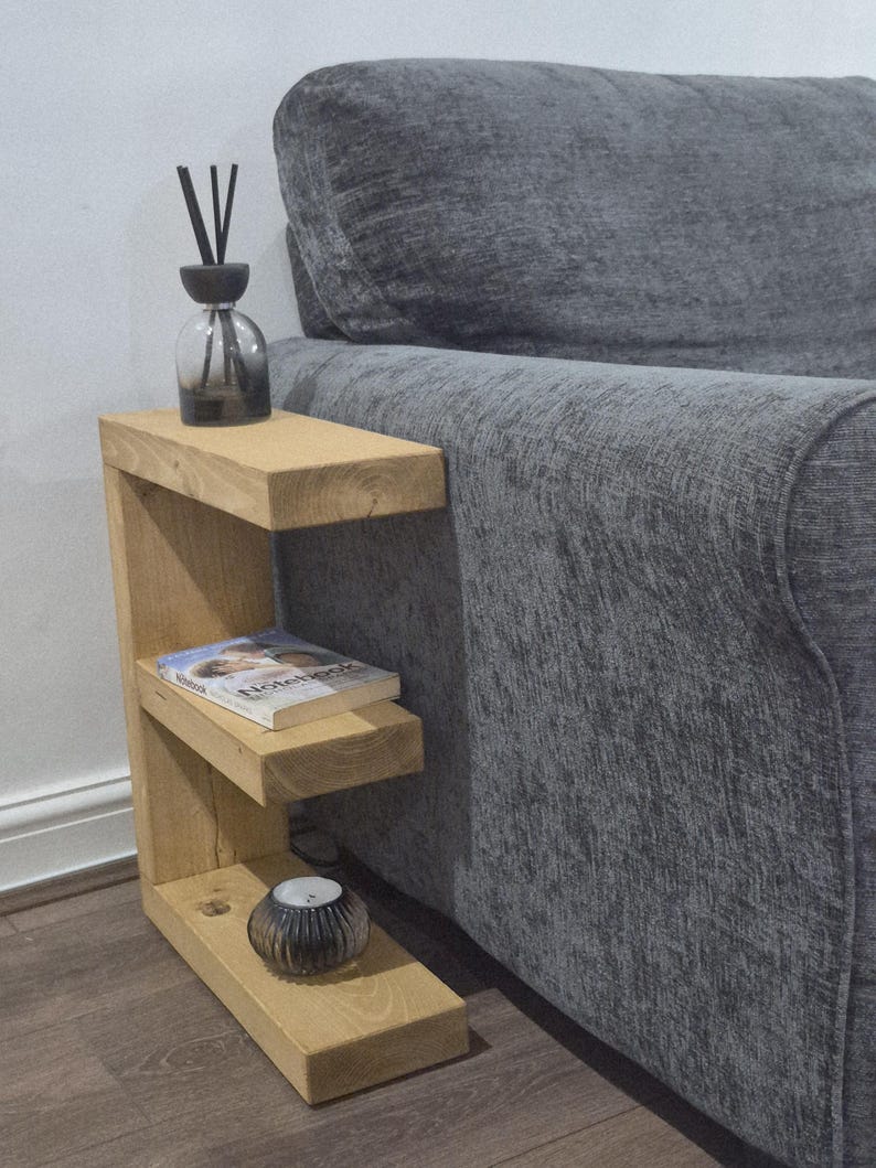 May include: A light-colored wooden side table with two shelves, positioned next to a gray sofa. The top shelf holds a glass diffuser with black reeds. A book and a small candle holder are on the lower shelves. The table is a functional piece of furniture.