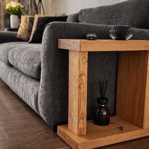 May include: A grey fabric sofa with a wooden side table. The table is made of natural wood and has a rectangular design. A black glass sphere and a small black object are on the table. A dark diffuser bottle is placed on the lower shelf.