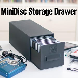 May include: A dark grey MiniDisc storage drawer with the text "MiniDisc Storage Drawer" above it. The drawer is open, revealing several MiniDiscs. A blue Sony MiniDisc player and other MiniDiscs are in the foreground.