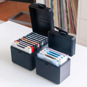May include: Two black rectangular storage cases with hinged lids, each containing multiple cassette tapes. The taller case holds a row of tapes, while the shorter case holds a smaller stack. The text "Long Live Physical Media" is embossed on the lids.