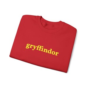 Può includere: Una maglietta rossa con la scritta "gryffindor" stampata in giallo. La maglietta è piegata e sembra essere fatta di un materiale morbido. Il testo è in un font sans-serif.