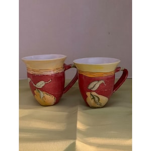 May include: Two red ceramic mugs with yellow and orange accents. The mugs feature a hand-painted design of pears and leaves.