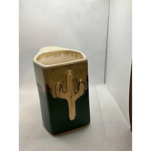 Puede incluir: Un jarrón rectangular de cerámica con un diseño de cactus. El jarrón tiene una base verde oscuro, un cactus dorado y un borde beige. El cactus está en relieve. El jarrón mide aproximadamente 15 cm de alto.