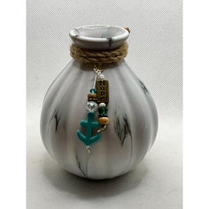 May include: A white ceramic vase with a marbled design and a rope accent around the neck. A charm with the word "Hope" and a turquoise anchor hangs from the vase.
