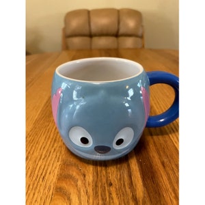 Puede incluir: Una taza de cer&aacute;mica azul con un personaje de dibujos animados de Stitch de Lilo & Stitch. La taza tiene un asa azul y orejas rosas.