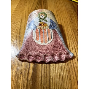 May include: A ceramic wall vase shaped like a woman in a pink dress with a blue bodice and a basket of flowers. The vase has a textured surface and is painted in various shades of brown, pink, blue, and green.