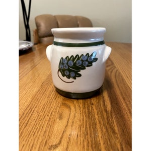May include: A small white ceramic jar with green and blue painted details. The jar has two small handles on the sides and a green band around the top and bottom. The jar features a painted design of a green leaf with blue berries.