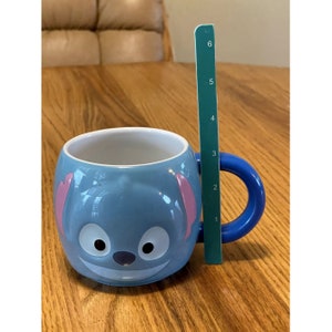 Puede incluir: Una taza de cer&aacute;mica azul con un dise&ntilde;o de personaje de Stitch. La taza tiene un asa azul y orejas rosas. La taza mide 10 cm de alto.