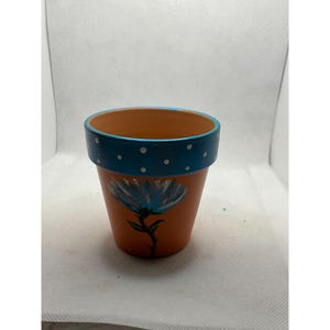 May include: A small, terracotta-coloured flower pot with a hand-painted blue flower design. The rim is painted blue with white polka dots. The pot is suitable for small plants and adds a decorative touch.