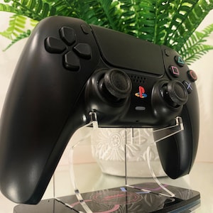 以下が含まれることがあります： 黒いPlayStation 5コントローラー。対称的なデザインで、タッチパッド、方向ボタン、アナログスティックを備えています。コントローラーは、カラフルなPlayStationロゴが付いた透明なアクリルスタンドに表示されています。背景には緑があります。