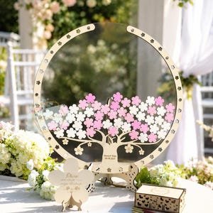 May include: A wooden wedding guest book alternative with a clear acrylic center, featuring a laser-cut tree design with pink and white flower accents. Includes a matching wooden box and pens, set on a white table with floral decorations.