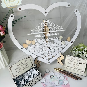 May include: White wooden heart-shaped guest book with a clear acrylic insert. The heart is decorated with a white crown and floral design. The text "Allison Martinez Quinceañera 09.24.2022" is printed on the heart. A small wooden box with a lid is filled with white wooden hearts. A sign reads "Please Sign a heart and drop it into the frame. Thank you for making my day special."