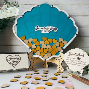 May include: A wooden seashell guest book with a teal blue interior. The seashell is on a wooden stand and has a sign that says "Guest Book" and "Please sign a and drop it into the frame so we will always remember our loved ones who came". The seashell is filled with small wooden hearts, some are gold and some are natural wood colour. The names "Joannie & Jerry" and the date "10.15.22" are written on the seashell.