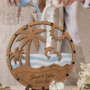 May include: A circular wooden sand ceremony frame featuring a cutout design of palm trees, a sun, and two dolphins. The frame is engraved with the names Steven & Helen and the date 15.05.2026. It rests on a decorative wooden stand.