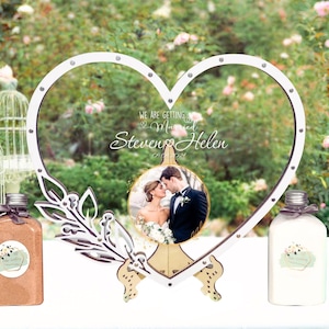 Könnte beinhalten: Weißer, herzförmiger Rahmen aus Holz mit einem Foto eines Paares darin. Der Rahmen ist mit einem floralen Design und dem Text "We are getting married Steven & Helen 01.01.2024" verziert.