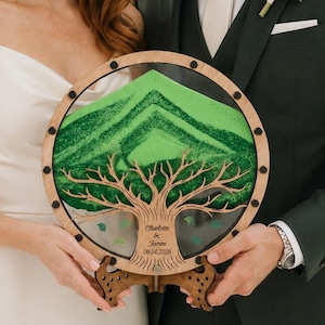 May include: A circular wooden sand ceremony frame featuring a laser-cut tree silhouette and green sand layered to form a mountain shape. The frame is engraved with the text Charlotte & James 06.14.2026 and includes a decorative wooden stand.
