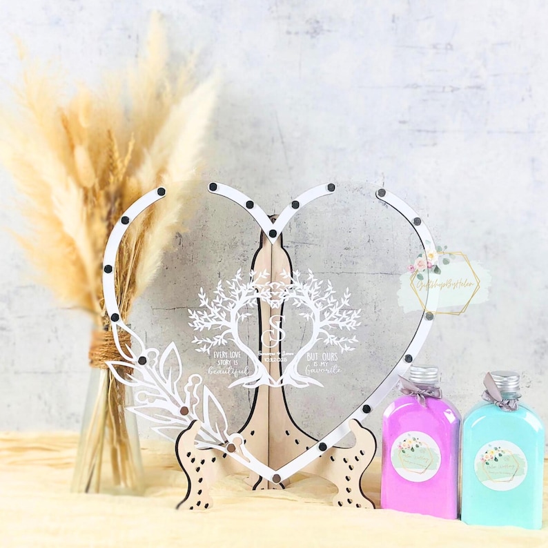 May include: A clear heart-shaped wedding sign with a white floral design and text. The sign is supported by a wooden stand. Two bottles of bath products in pink and blue are in front of the sign. A vase of dried flowers is on the left.