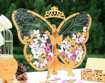 Puede incluir: Un libro de visitas de mariposa de madera con un diseño de alas de acrílico transparente. La mariposa está decorada con una corona y tiene un letrero que dice "Helen Sweet 16". El libro de visitas viene con una caja de pequeñas mariposas de madera que los invitados pueden firmar y colocar en las alas.